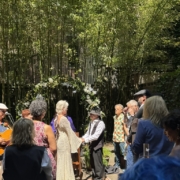 Couple in vintage clothing exchange vows beneath a flower arch in a sunlit garden ceremony.