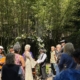 Couple in vintage clothing exchange vows beneath a flower arch in a sunlit garden ceremony.
